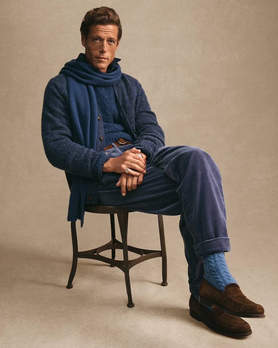 Man seated wearing blue knitwear and matching scarf with brown loafers.