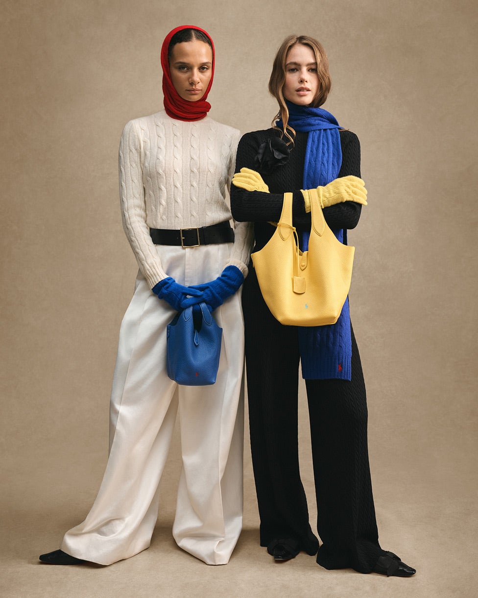 Due donne in piedi: una con una maglia color panna, copricapo rosso e guanti blu; l’altra con una maglia nera, sciarpa blu e borsa gialla.