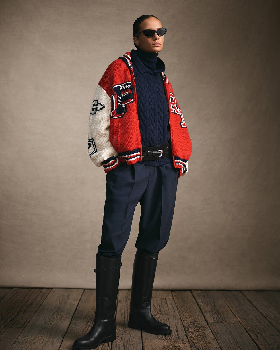 Donna in piedi con una giacca in stile college rossa e bianca, maglia e pantaloni blu navy e stivali alti neri.