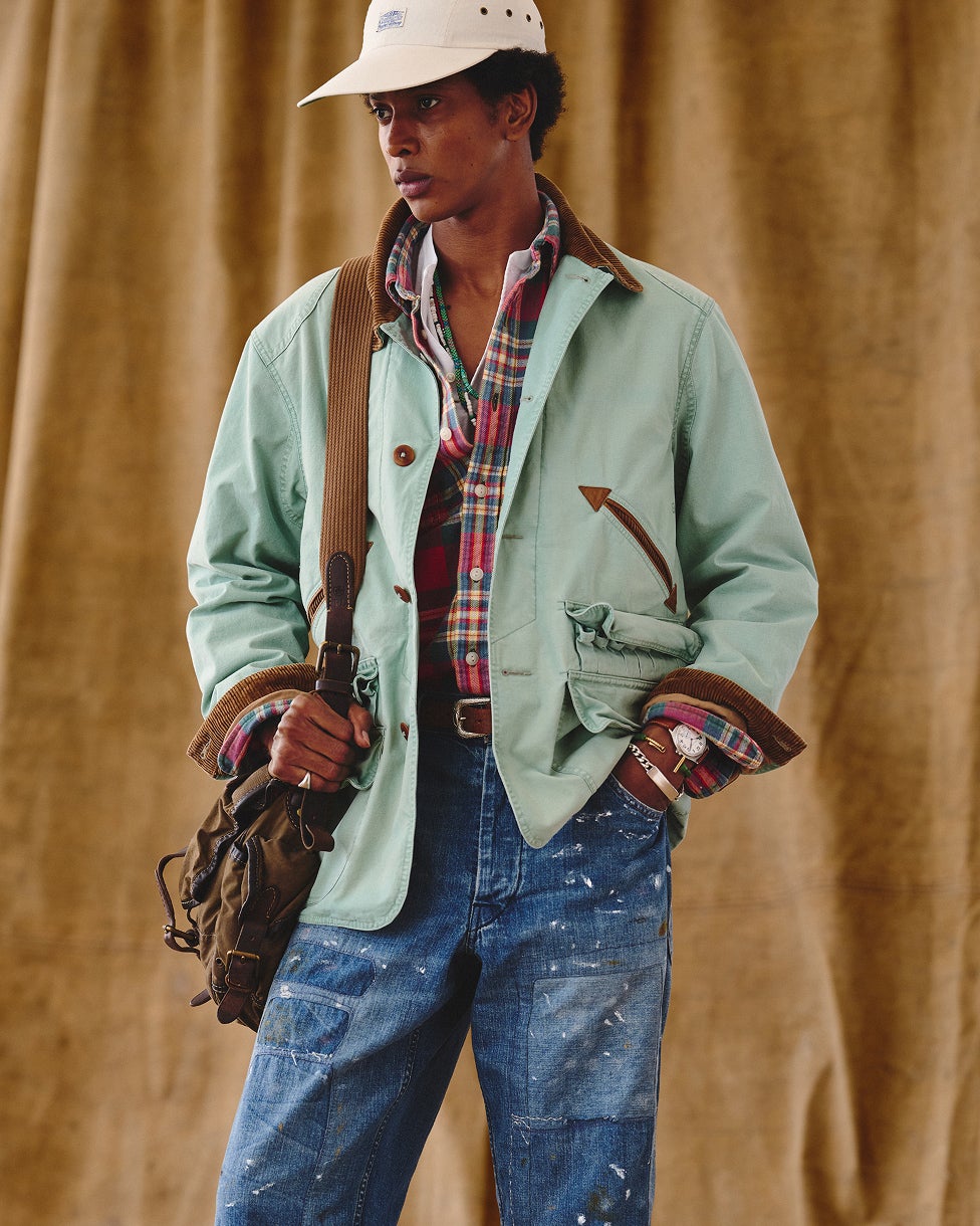Uomo che indossa una giacca verde sopra una camicia scozzese, jeans blu, un cappellino color panna e una borsa.