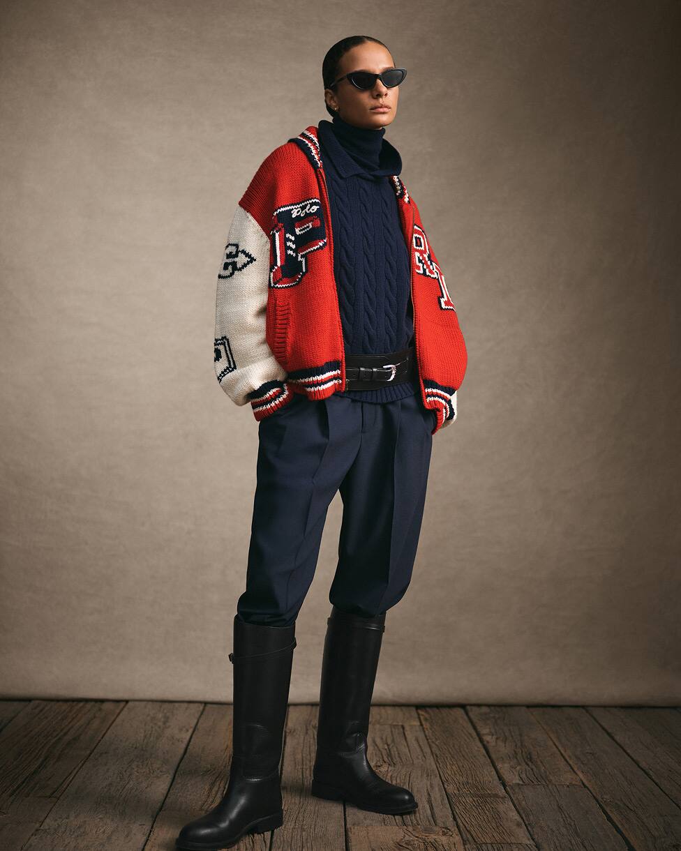 Donna in piedi con una giacca in stile college rossa e bianca, maglia e pantaloni blu navy e stivali alti neri.